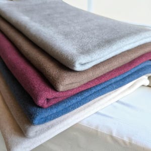 Op de afbeelding: Een stapel gevouwen stukken stof in verschillende effen kleuren, waaronder lichtgrijs, bruin, dieprood, donkerblauw en crème. Elk stuk stof heeft een zachte, vervilte textuur met zichtbare vezels en strakke, rechte randen die netjes op elkaar liggen.