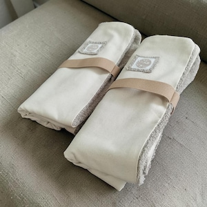 May include: Two folded rectangular towels featuring a smooth off-white fabric on one side and a textured beige terry cloth on the other. Each towel is secured with a tan elastic band and features a small square patch with a circular logo design.