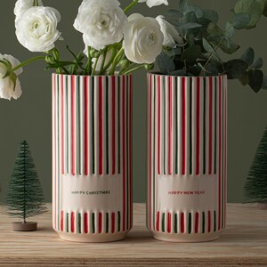 May include: Two white ceramic vases with red and green stripes. The vase on the left says "Happy Christmas" and the vase on the right says "Happy New Year". The vases are filled with white flowers and green leaves.