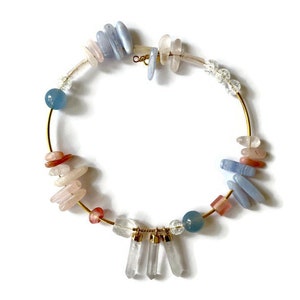 Può includere: Un girocollo in filo d'oro con una varietà di pietre blu, rosa e bianche. Le pietre sono disposte in modo casuale attorno al cerchio.