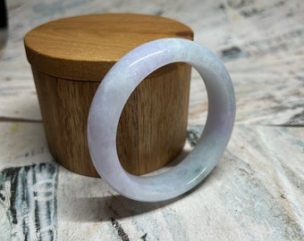 May include: A light lavender jade bangle bracelet with a smooth, polished finish. The bangle is displayed on a wooden box.