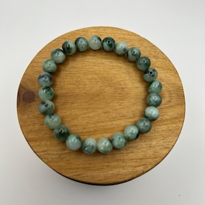 Op de afbeelding: Een armband met ronde, gepolijste kralen in groen- en grijstinten. De armband wordt gepresenteerd op een ronde, houten ondergrond, die de natuurlijke kleur- en textuurvariaties van de steen benadrukt. De kralen hebben een diameter van ongeveer 1,3 cm.