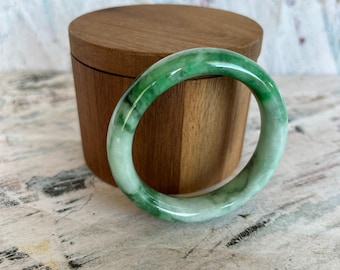 May include: A jade bangle bracelet with a mix of light and dark green hues. The bracelet is circular and sits in front of a wooden box with a lid. The background is a light-colored surface with dark markings.