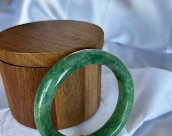 May include: A green jade bangle bracelet with a red string tied around it. The bracelet is sitting on a wooden box.