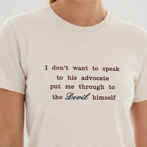 May include: A cream-colored short-sleeve t-shirt with a crew neckline. The shirt features dark red text that reads: I don't want to speak to his advocate put me through to the Devil himself, with the word Devil written in a dark blue script font.