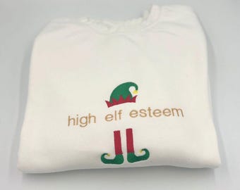 May include: White sweatshirt with a green elf graphic and the text "high elf esteem" in brown embroidery.