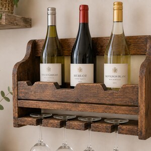 May include: A rustic, dark-stained wooden wine rack holding three wine bottles. The bottles are labeled Pinot Grigio, Merlot, and Sauvignon Blanc. The rack also has space for wine glasses below.