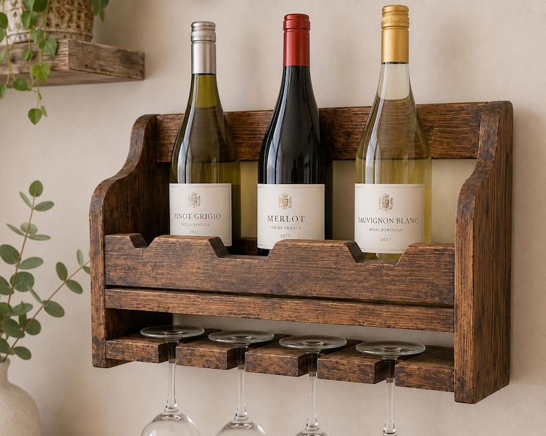 May include: A rustic, dark-stained wooden wine rack holding three wine bottles. The bottles are labeled Pinot Grigio, Merlot, and Sauvignon Blanc. The rack also has space for wine glasses below.