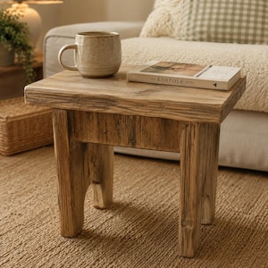 May include: A small, weathered wooden side table with a natural finish. A beige ceramic mug and a book titled "The Kinfolk Home" sit on the tabletop. The table has a square top and four sturdy legs, ideal for a living room.