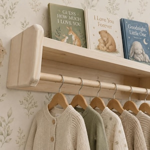 May include: A light-coloured wooden wall shelf with a horizontal rod for hanging clothes. The shelf holds three children's books titled Guess How Much I Love You, I Love You Forever, and Goodnight, Little One. Six wooden hangers hold various sweaters.