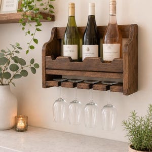 May include: A dark wood wine rack holding three wine bottles and four wine glasses. The rack is mounted on a white wall above a countertop. The bottles are labeled "Sancerre", "Bourgogne", and "The Dreaming Angel".