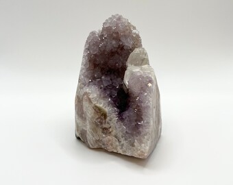 Könnte beinhalten: Eine natürliche Amethyst-Geode mit einer Ansammlung von tiefvioletten, kristallinen Spitzen, die sich in einer zentralen Höhle konzentrieren. Das Äußere des Steins zeigt eine raue, hellgraue und weiße Gesteinsformation mit Texturen.