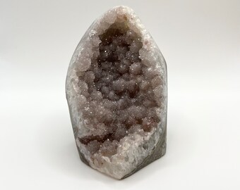 Könnte beinhalten: Eine vertikale Amethyst-Geode mit einem hohlen Zentrum, das mit kleinen, funkelnden, tiefvioletten Kristallformationen gefüllt ist. Die Außenkanten des Steins bestehen aus einer glatten, hellen Mineralkruste, die das dunkle, texturierte Innere einrahmt.