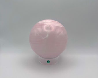 Könnte beinhalten: Ein glatter, blassrosa kugelförmiger Stein mit dezenter horizontaler Bänderung und glänzender Oberfläche. Die Kugel ruht auf einem durchsichtigen, runden Kunststoffständer, der einen kleinen grünen Aufkleber mit den schwarzen Buchstaben MZ trägt.
