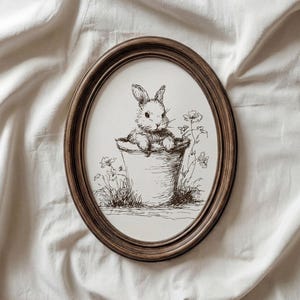 Puede incluir: Un dibujo a pluma y tinta enmarcado de forma ovalada de un conejito en una maceta. El conejito está dibujado con detalle con una apariencia esponjosa. El marco es marrón con aspecto de veta de madera. El fondo es una sábana blanca.