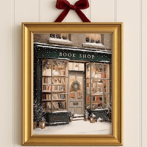 May include: A framed print of a cozy bookstore in winter. The gold frame surrounds a scene with snow-covered ground, a wreath on the door, and the text "BOOK SHOP" above the windows. A red bow hangs above the frame.