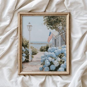 May include: A framed print of a coastal scene with a house, American flag, and blue hydrangeas. The artwork features a light post, a path, and a view of the ocean. The frame is a light wood color.