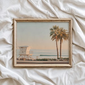 Puede incluir: Impresión enmarcada de una escena costera con una torre de salvavidas, palmeras y olas del océano. La obra de arte tiene un marco de madera marrón claro y una paleta de colores pastel serena.