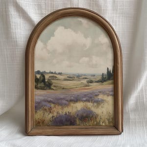 Puede incluir: Pintura de paisaje enmarcada, con un campo de lavanda en primer plano. El marco es de madera marrón claro. El cielo está nublado, y el fondo muestra colinas y árboles. La obra de arte evoca serenidad.