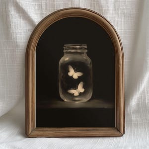 Pode incluir: Pintura emoldurada com um frasco de vidro transparente com duas borboletas brancas no interior, contra um fundo escuro. A moldura é castanho-claro. A obra de arte tem uma qualidade suave e etérea.