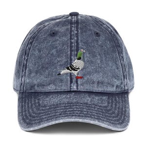 Peut inclure: Une casquette de baseball bleu marine délavée à l'acide avec un pigeon brodé sur le panneau avant. Le pigeon est représenté dans des tons de gris, blanc et noir avec une tache verte au cou et un petit détail rouge près de ses pattes.