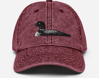 Può includere: Un cappellino da baseball bordeaux con un disegno ricamato di un colimbo. Il colimbo è raffigurato in bianco e nero, con il corpo e la testa dell'uccello ben visibili. Il cappellino ha una visiera curva e un bottone sulla sommità.