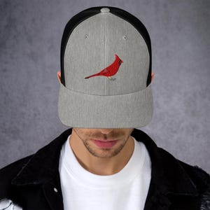 Op de afbeelding: Een grijze en zwarte truckerpet met een rood kardinaalvogelontwerp. Het voorpaneel is grijs met de kardinaal erop geborduurd. De achterkant van de pet is zwart mesh en de klep is grijs.
