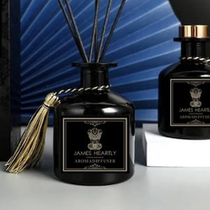 May include: Two black glass bottles with gold accents and labels that read "JAMES HEARTLY AROMADIFFUSER". One bottle has a gold tassel and black reeds sticking out of the top. The bottles are sitting on a white surface.