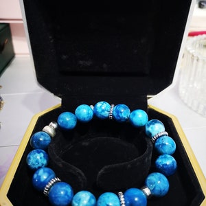Può includere: Un braccialetto di pietre blu con accenti argentati in una scatola di velluto nero. Il braccialetto è fatto di perline rotonde con un motivo marmoreo.