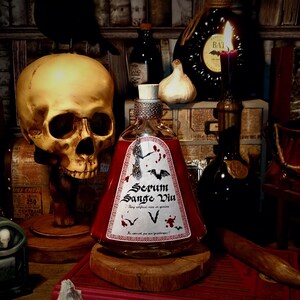 Può includere: Una bottiglia di vetro con un liquido rosso e un'etichetta che recita "Serum Sangue Vin" in caratteri neri. La bottiglia è appoggiata su una superficie di legno con un teschio e altri oggetti sullo sfondo.