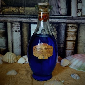 Op de afbeelding: Een glazen fles met een kurkstop gevuld met een blauwe vloeistof. De fles heeft een etiket met de tekst "Larmes de Sirenes". De fles staat op een zanderige ondergrond met schelpen.