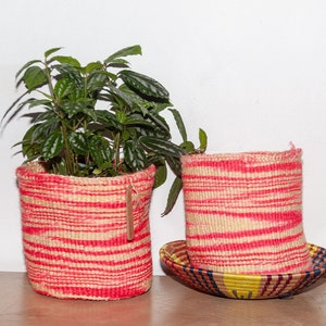 Puede incluir: Dos cestas tejidas para plantas con rayas horizontales rojas y beige. Una cesta contiene una planta verde con hojas. La otra se encuentra sobre un platillo tejido a juego. Las cestas están hechas de fibras naturales y están sobre una superficie de madera.