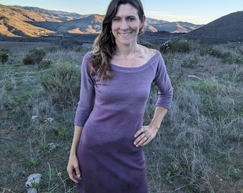 May include: A woman wearing a purple ombre dress with a boat neck and three-quarter sleeves. She is standing in a field with hills in the background.