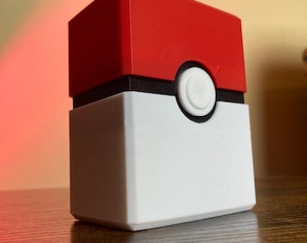 Peut inclure: Une boîte de rangement pour cartes Pokemon imprimée en 3D, de couleur rouge, blanche et noire. La boîte a un motif de Poké Ball sur le devant.