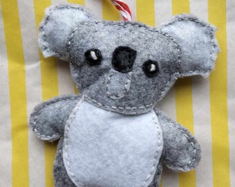 Può includere: Un ornamento a forma di koala in feltro grigio con pancia in feltro bianco e occhi ricamati neri. L'ornamento è appeso a uno spago rosso e bianco.
