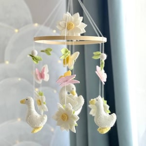 Puede incluir: Móvil para bebé hecho a mano con cisnes, flores, mariposas y hojas de fieltro en colores pastel. El móvil está suspendido de un aro de madera y presenta delicados detalles de hilo, creando una decoración infantil.