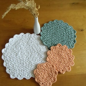 Könnte beinhalten: Eine Sammlung handgefertigter Häkelartikel in verschiedenen Farben. Das Bild zeigt eine große weiße Tischdecke, eine mintgrüne Tischdecke und zwei pfirsichfarbene Untersetzer. Eine kleine weiße Vase mit Trockenblumen befindet sich im Hintergrund. Die Artikel liegen auf einer Holzoberfläche.