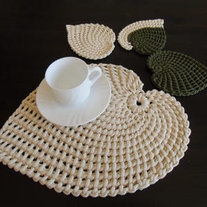 Könnte beinhalten: Ein Set gehäkelter Untersetzer in Blattform in Beige und Grün, mit einer weißen Untertasse und Tasse auf einem größeren beigefarbenen gehäkelten Untersetzer.