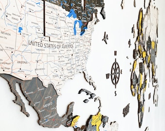 May include: A wooden world map with a 3D effect. The map is painted in shades of gray and black, with yellow accents. The map is mounted on a white wall. The map includes a compass and the text "UNITED STATES OF AMERICA".