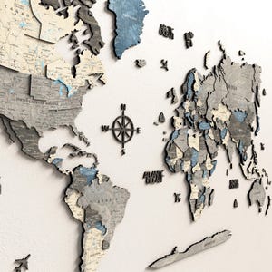 Op de afbeelding: Een 3D houten wereldkaart in tinten grijs, blauw en beige, gemonteerd op een wit oppervlak. De kaart toont gedetailleerde contouren en labels van landen, met een kompasroos in het midden. De Atlantische Oceaan en Antarctica zijn gelabeld.