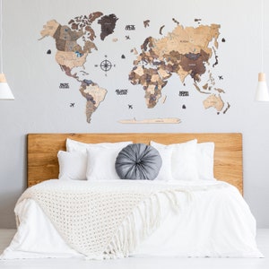 Puede incluir: Un mapa del mundo de madera con un acabado marr&oacute;n claro, que incluye una br&uacute;jula y los nombres de los oc&eacute;ans. El mapa est&aacute; montado en una pared blanca sobre una cabecera de madera con ropa de cama blanca y una almohada gris.