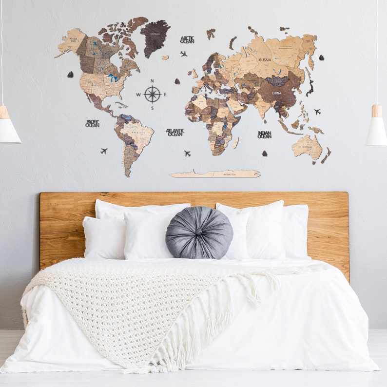 Puede incluir: Un mapa del mundo de madera con un acabado marr&oacute;n claro, que incluye una br&uacute;jula y los nombres de los oc&eacute;ans. El mapa est&aacute; montado en una pared blanca sobre una cabecera de madera con ropa de cama blanca y una almohada gris.