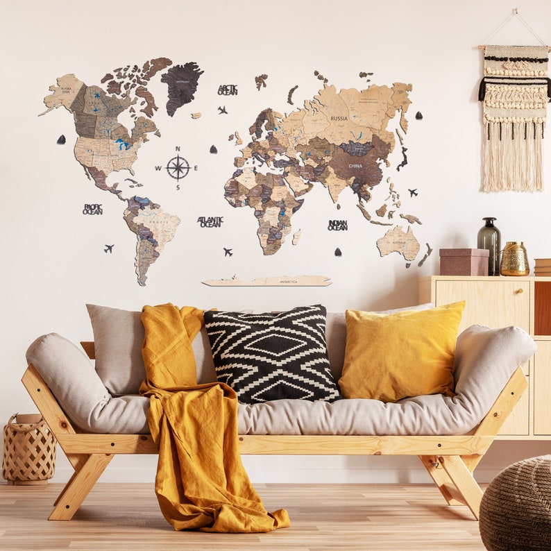 Puede incluir: Un mapa del mundo de madera con un acabado marr&oacute;n claro, que incluye una br&uacute;jula y etiquetas de los oc&eacute;anos. El mapa est&aacute; montado en una pared blanca sobre un sof&aacute; marr&oacute;n claro con almohadas de patrones amarillos y negros.