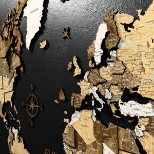 Puede incluir: Un detallado mapa del mundo de madera con fondo negro. El mapa presenta países en varios tonos de marrón y blanco, con nombres de ciudades y una rosa de los vientos. Se incluyen detalles decorativos de avión y velero.