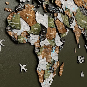 Peut inclure: Une carte du monde en bois détaillée, avec des pays de différentes couleurs, dont le marron, le vert et le blanc. La carte présente les noms des pays et est montée sur un fond vert foncé. Des accents décoratifs d'avion et de voilier sont inclus.