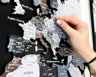 May include: A black and white world map with a brown textured surface. The map is mounted on a black background. A hand is placing a pin on the map in the area of Ukraine.