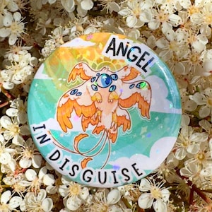 May include: A circular badge featuring the words "ANGEL IN DISGUISE" and a cartoon illustration of a multi-eyed, winged creature. The badge has a light blue, green, and yellow background, set against a backdrop of white flowers.