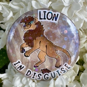 May include: A circular badge featuring a cartoon lion with a brown mane and body. The text "LION IN DISGUISE" is displayed in white. The background has a glitter effect. The badge is set against a backdrop of white flowers.