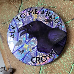 Puede incluir: Un botón redondo con un fondo holográfico y una ilustración de cuervo morado. El botón dice "TALK TO ME ABOUT CROWS" en texto negro. El botón está colocado sobre una superficie marrón texturizada, con una llave en primer plano.
