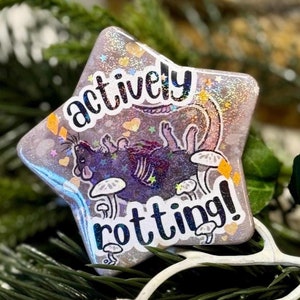May include: A glittery star-shaped resin pin with a black and white illustration of a rat on mushrooms. The text "actively rotting!" is printed on the pin.